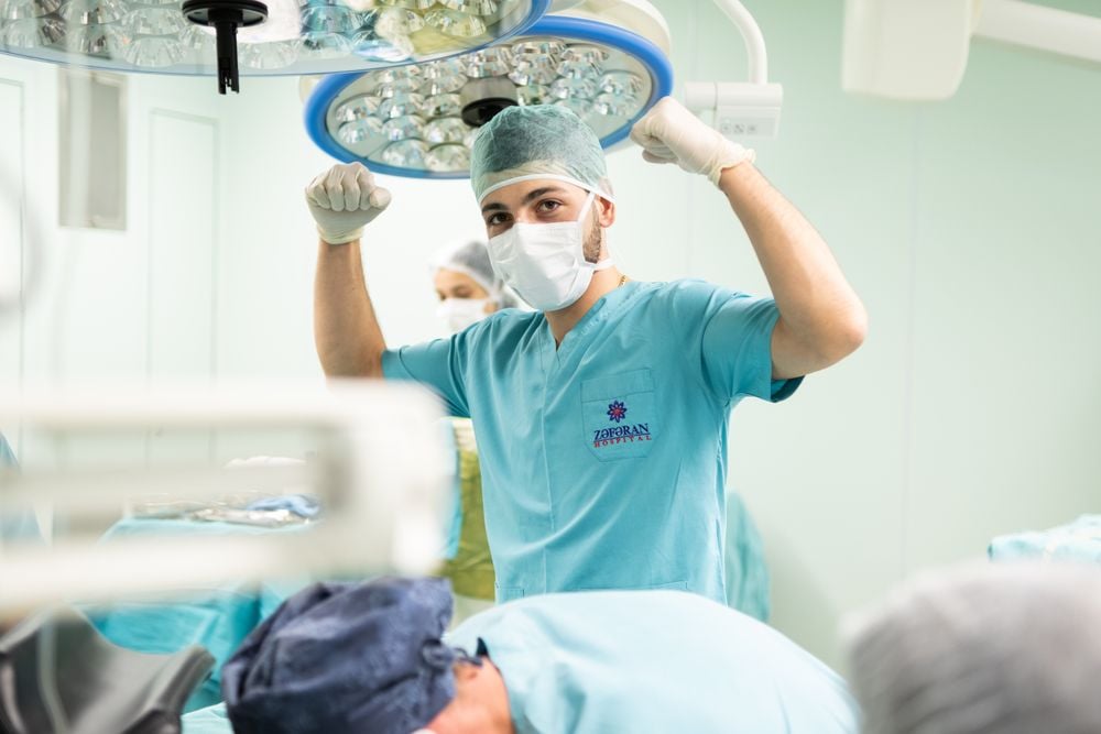 A surgeon in scrubs and mask flexes his arms playfully in an operating room, while another medical staff member and a patient lying on the table are visible in the background.