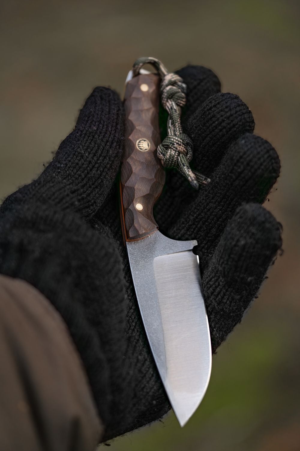 A person wearing a black glove holds a small fixed-blade knife with a textured wooden handle and a lanyard attached, against a blurred outdoor background.