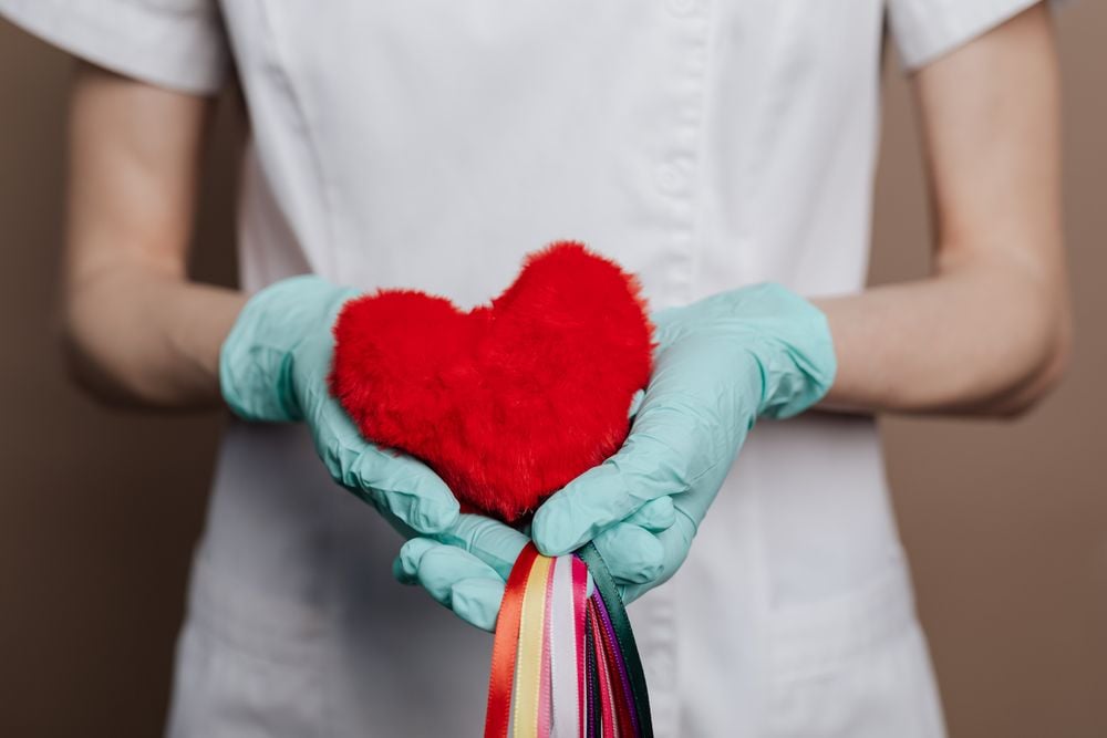 A person in a white uniform and blue gloves holds a red, plush heart with colorful ribbons hanging from it.