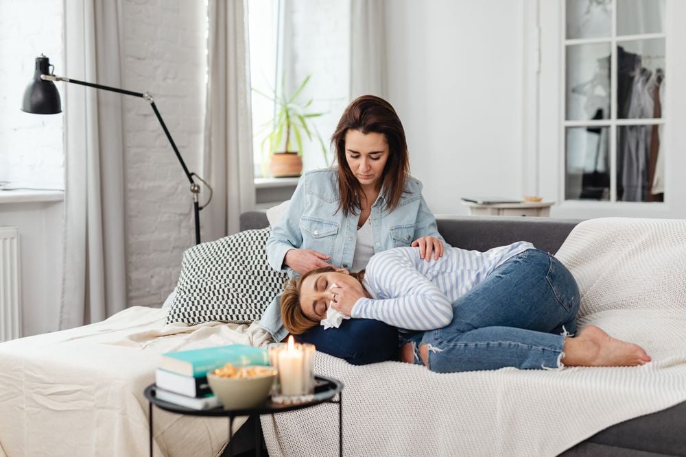 A woman comforts another woman lying on a couch, who appears to be upset and holding a tissue. The scene is cozy, with a candle, books, and snacks on a table in a softly lit living room.
