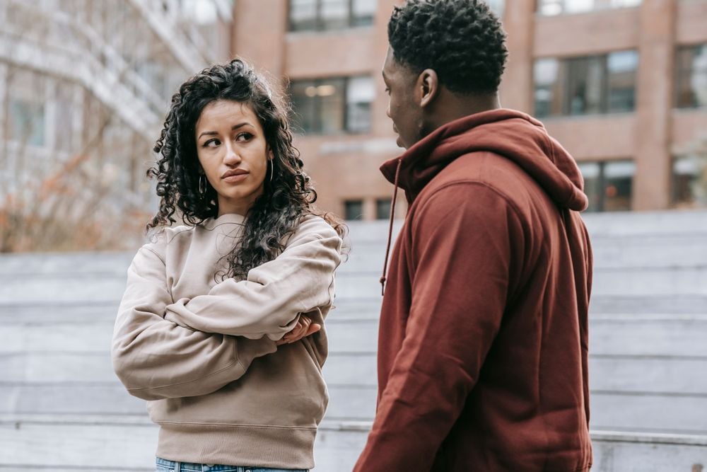 A woman with curly hair stands with arms crossed, looking at a man in a red hoodie. They appear to be having a serious conversation outdoors near steps and a building.