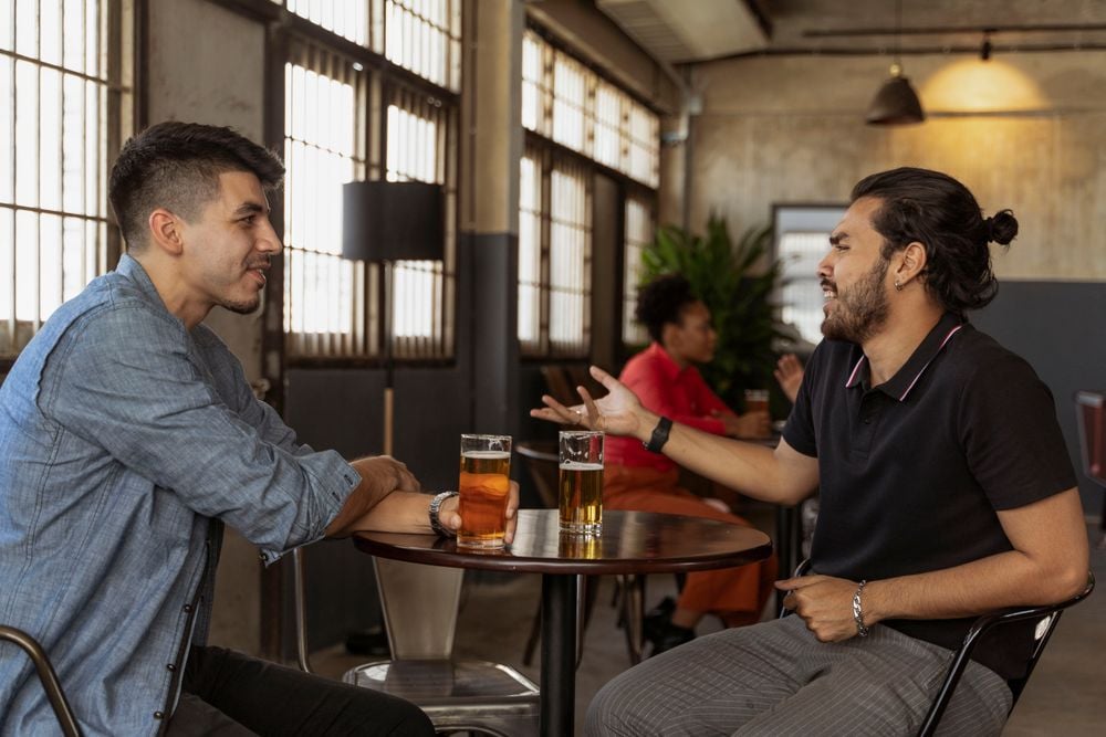 Two men sit at a table in a bar, talking with drinks in front of them. One gestures expressively while the other listens. In the background, two people talk at another table.