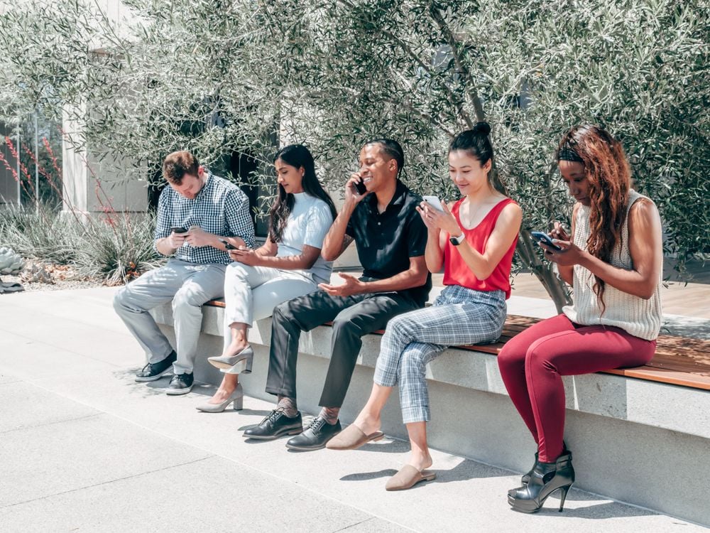 Five people sit on a bench outdoors, each focused on their smartphones. Trees and plants are in the background, and the weather appears sunny.