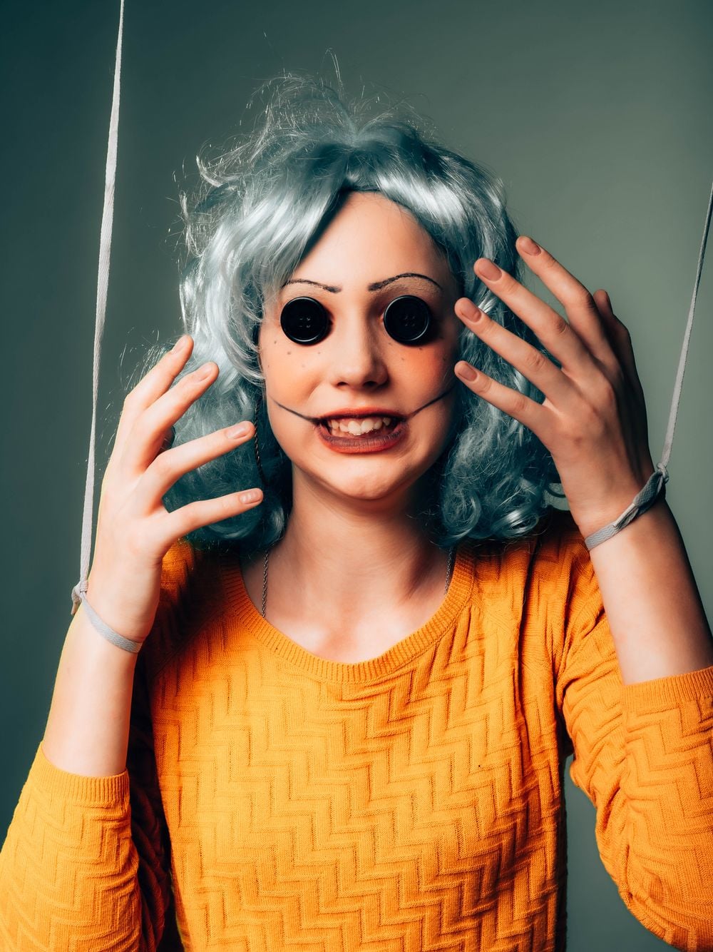 Person dressed as a doll with button eyes, drawn-on mouth lines, a blue wig, and an orange sweater, hands raised to their head. Strings tied to wrists suggest a marionette puppet.