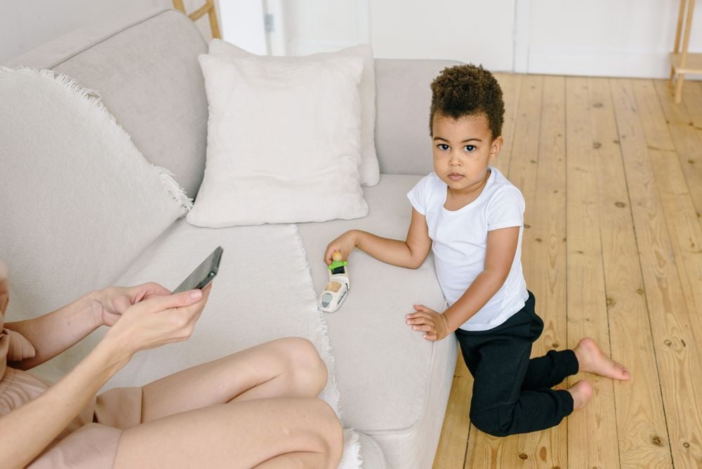 A young child kneels on a wooden floor next to a sofa, holding a toy. An adult, partially visible, sits on the sofa using a smartphone.