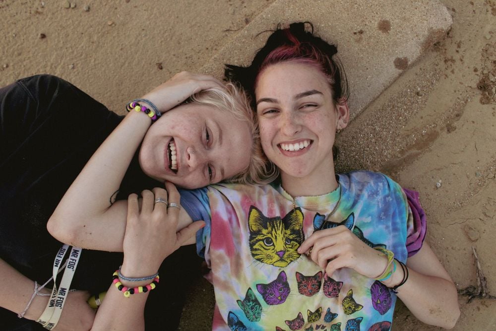 Two smiling young people lie on sandy ground, one wearing a colorful cat-themed shirt, both holding hands and wearing friendship bracelets.