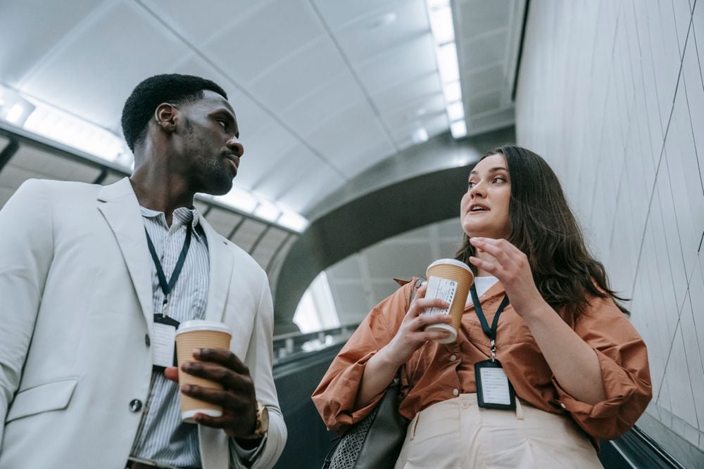 Two professionals wearing ID badges talk while holding coffee cups, standing on an escalator in a modern building with a curved ceiling.