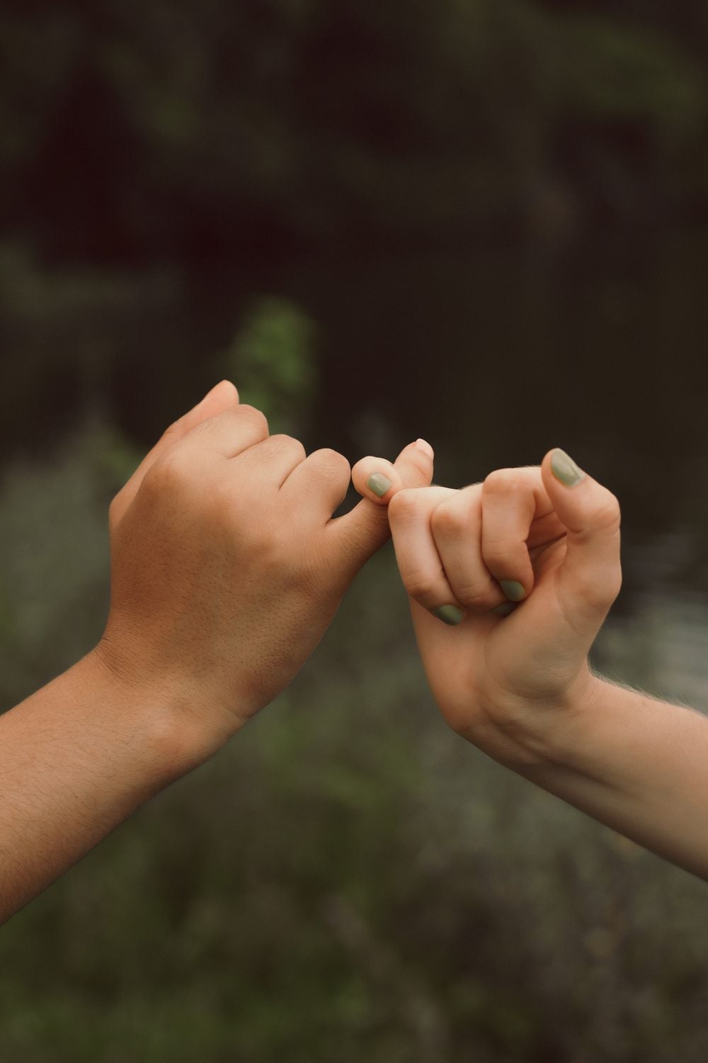 Two hands making a pinky promise against a blurred outdoor background. One hand has a darker skin tone and the other a lighter skin tone, both with short nails.