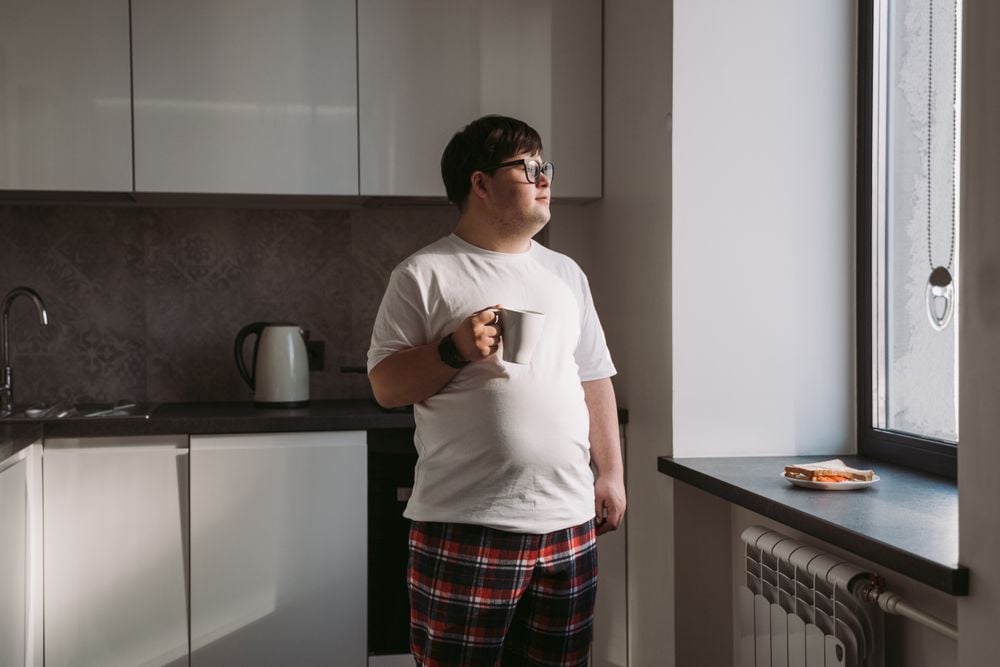 A man in pajamas stands in a kitchen holding a mug, looking out the window. Sunlight comes through the window, and a plate with food sits on the windowsill.