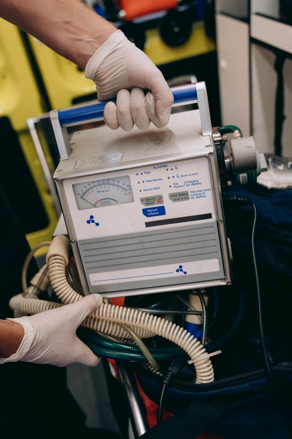 A person wearing white gloves holds a portable medical device with dials, buttons, and hoses, likely used for respiratory support or monitoring.