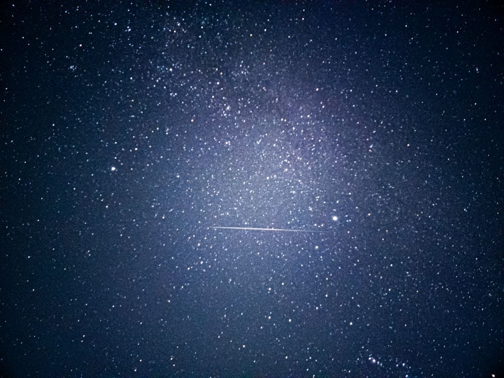 A clear night sky filled with stars, featuring a single bright meteor streaking horizontally across the center.