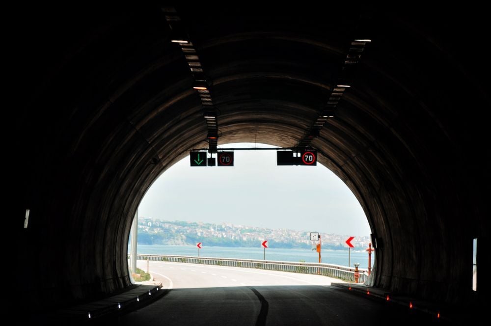 View from inside a tunnel looking out toward a road curving by the water. Speed limit signs and an arrow signal hang above the tunnel exit. Distant hills and sky are visible.