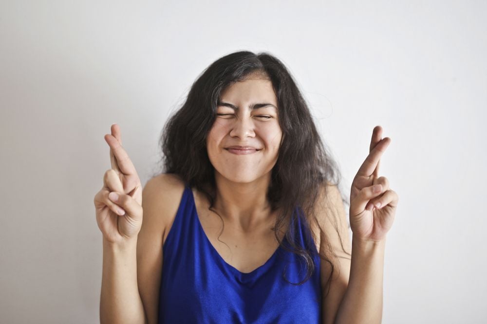 Woman with long dark hair wearing a blue tank top smiles with eyes closed and both hands raised, fingers crossed, standing against a plain white background.
