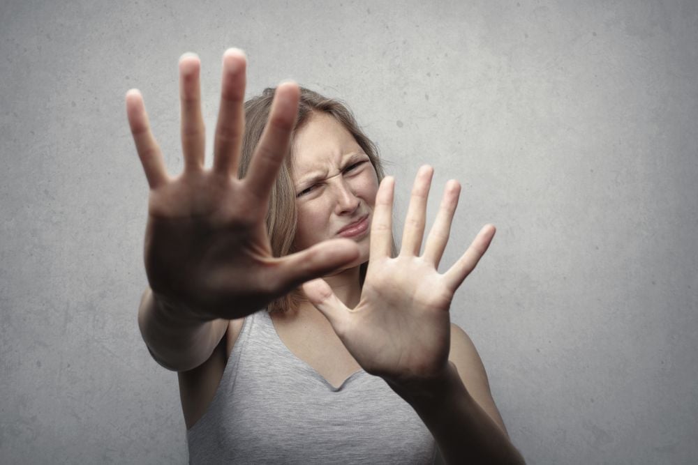 A woman in a gray tank top holds her hands up in front of her, palms facing outward, as if to block or reject something, with a distressed expression on her face against a plain gray background.