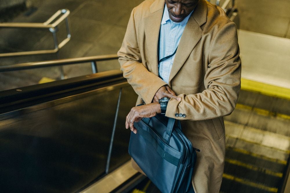 A man in a tan coat rides an escalator while looking at his wristwatch and holding a black briefcase.