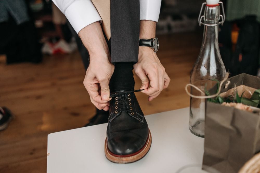 A person in dress pants and a watch is tying the laces of a black dress shoe while resting their foot on a white surface. A glass bottle and a brown paper bag are nearby on a wooden floor.