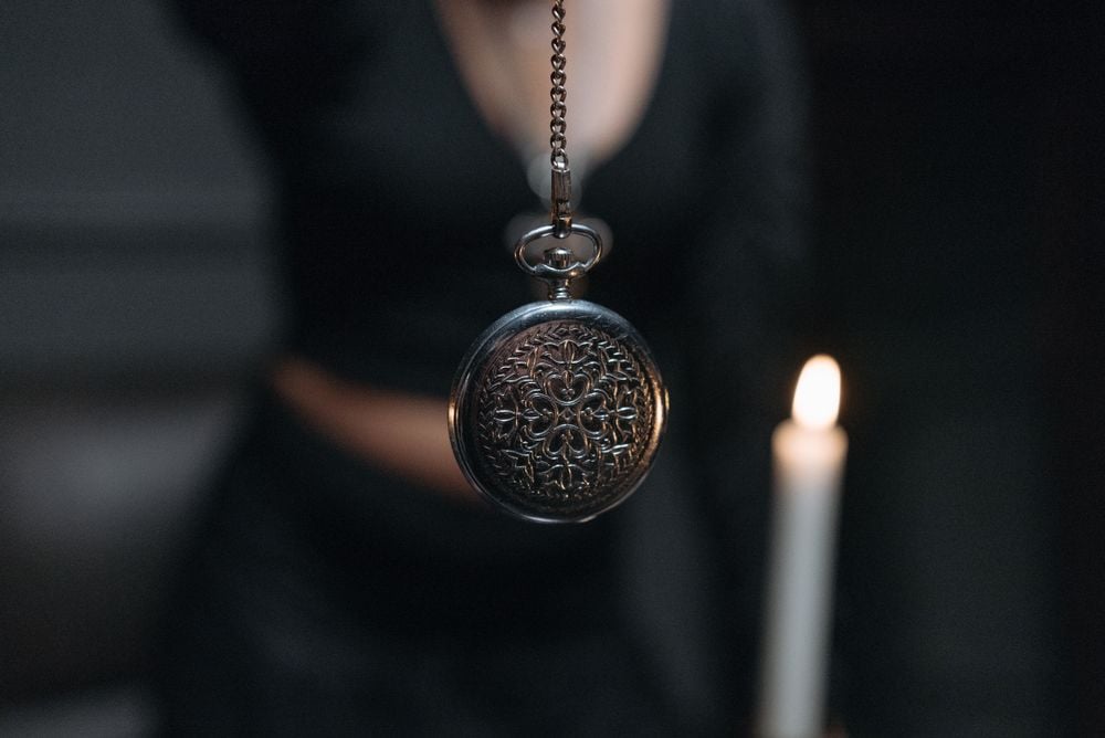 A person holds a detailed pocket watch by its chain in front of them. A lit candle is visible in the background, creating a mysterious, dimly lit atmosphere.