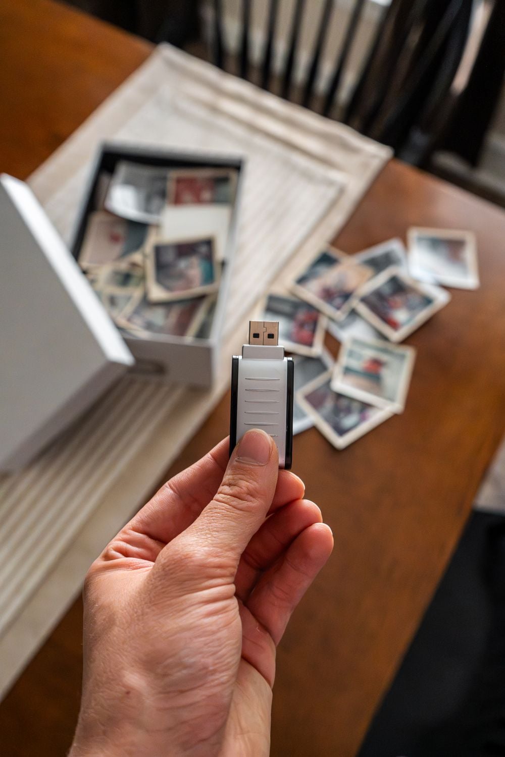 A hand holds a USB flash drive over a table with scattered old photographs and a box filled with more vintage photos in the background.