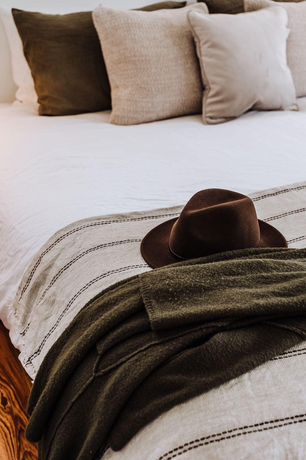 A brown hat rests on a striped blanket at the foot of a neatly made bed with earth-toned pillows and a cozy, inviting atmosphere.
