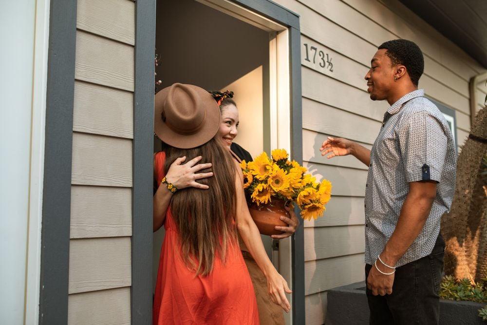 A woman in a red dress hugs another woman holding a bouquet of sunflowers at a front door, while a man stands nearby, smiling. The house number 173½ is visible on the siding.