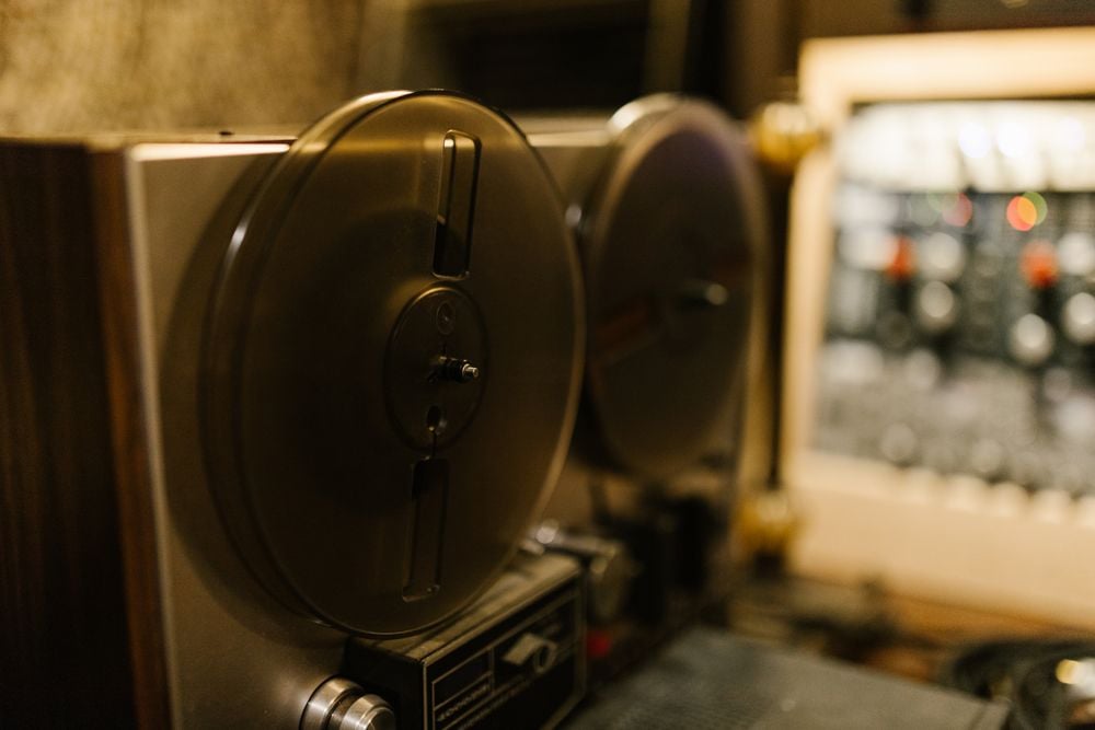 Close-up of a vintage reel-to-reel tape recorder with two metal reels; blurred audio equipment is visible in the background.