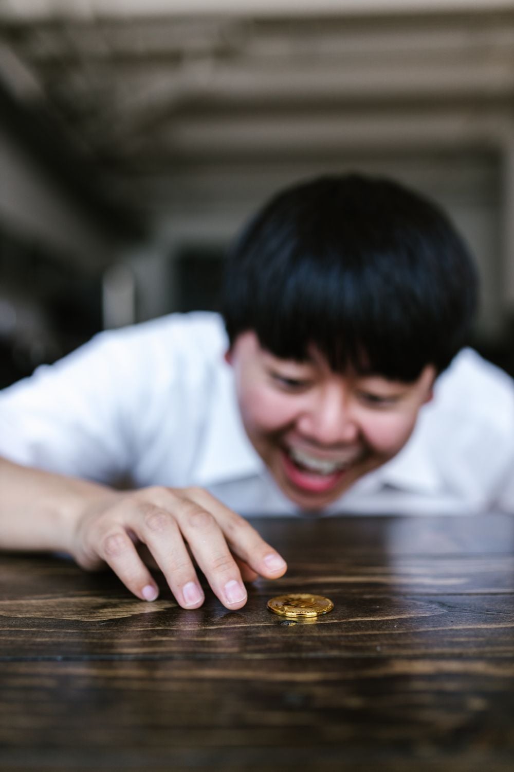 A person with short dark hair smiles excitedly while reaching for a gold coin on a wooden table.