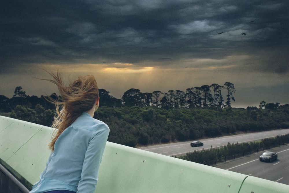 A person with long hair blowing in the wind leans over a green railing, looking at a dark, cloudy sky above a highway with a few cars and trees. Sunlight breaks through the clouds in the distance.