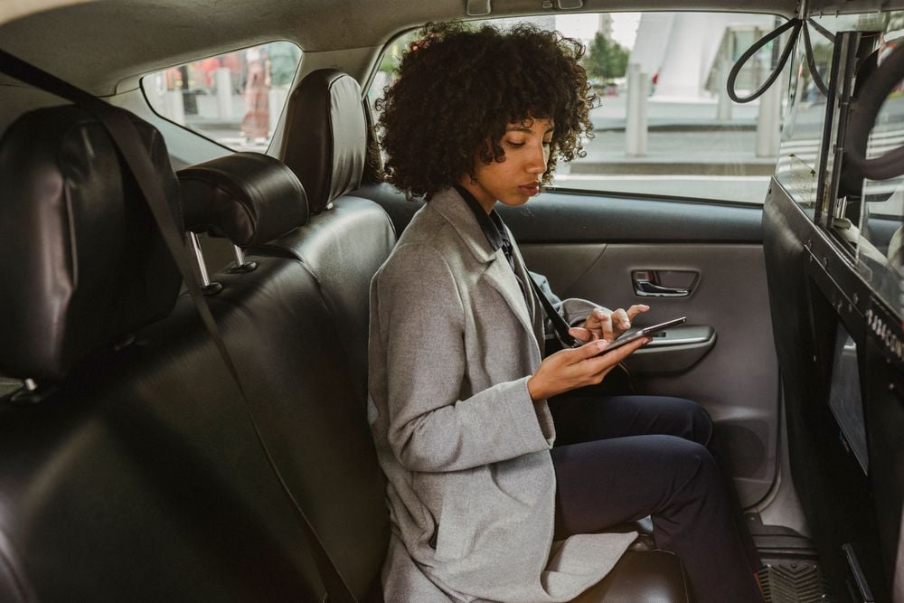 A person with curly hair, wearing a gray coat and dark pants, sits in the backseat of a car, looking at their phone.