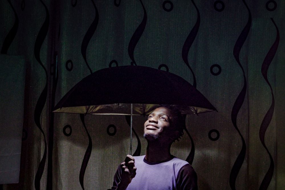A person stands indoors holding an open umbrella and looking upward, smiling. The background features a curtain with a wavy black pattern and circular designs.
