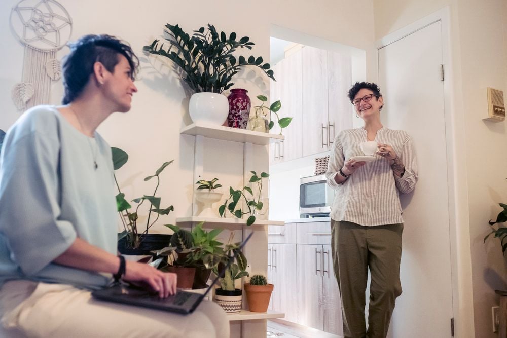 Two people talk and smile in a cozy room with many plants. One sits with a laptop, while the other stands in the doorway holding a mug. The space is warmly lit and inviting.