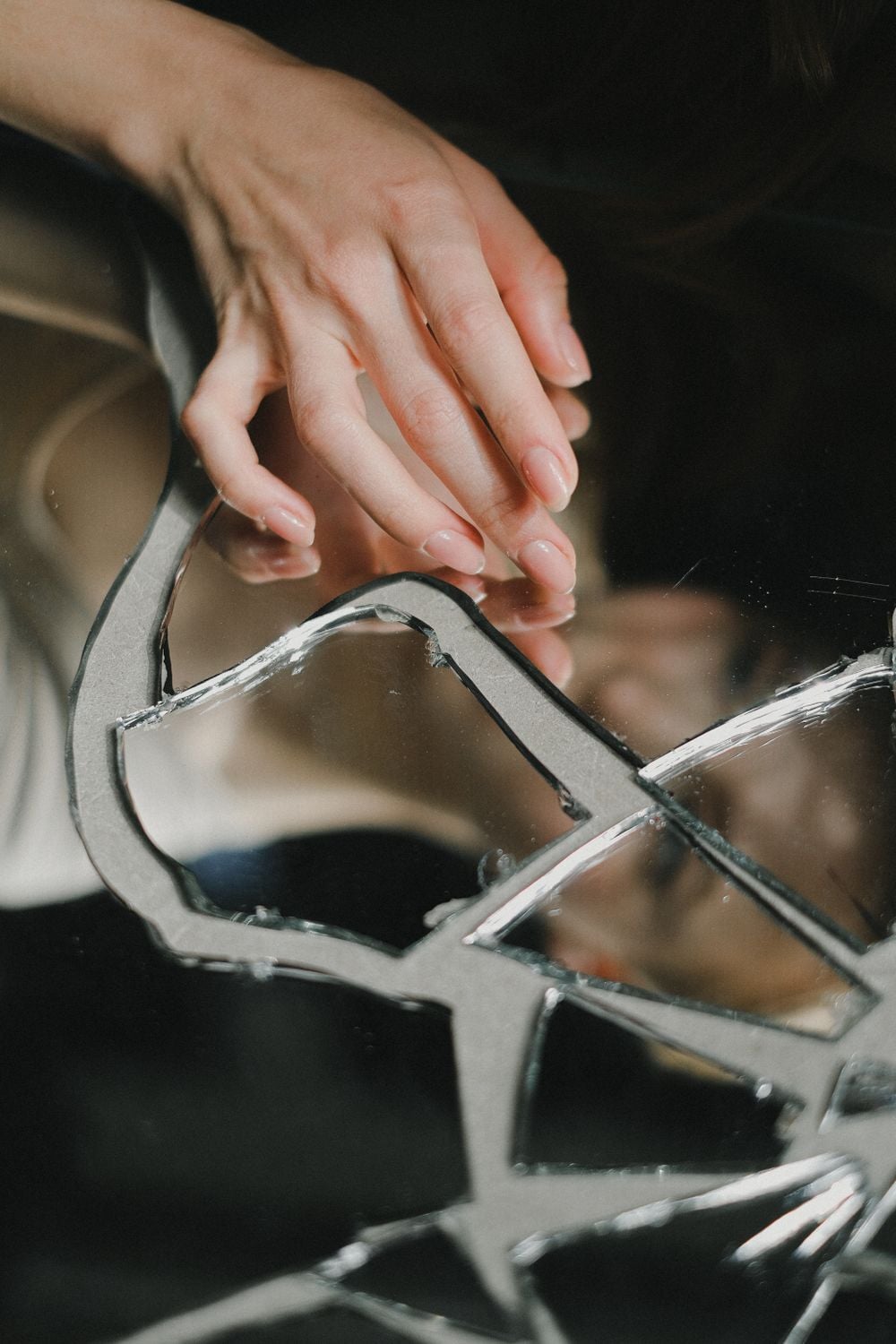 A hand rests gently on a cracked mirror, with the reflection of the hand and part of a face visible among the broken shards.