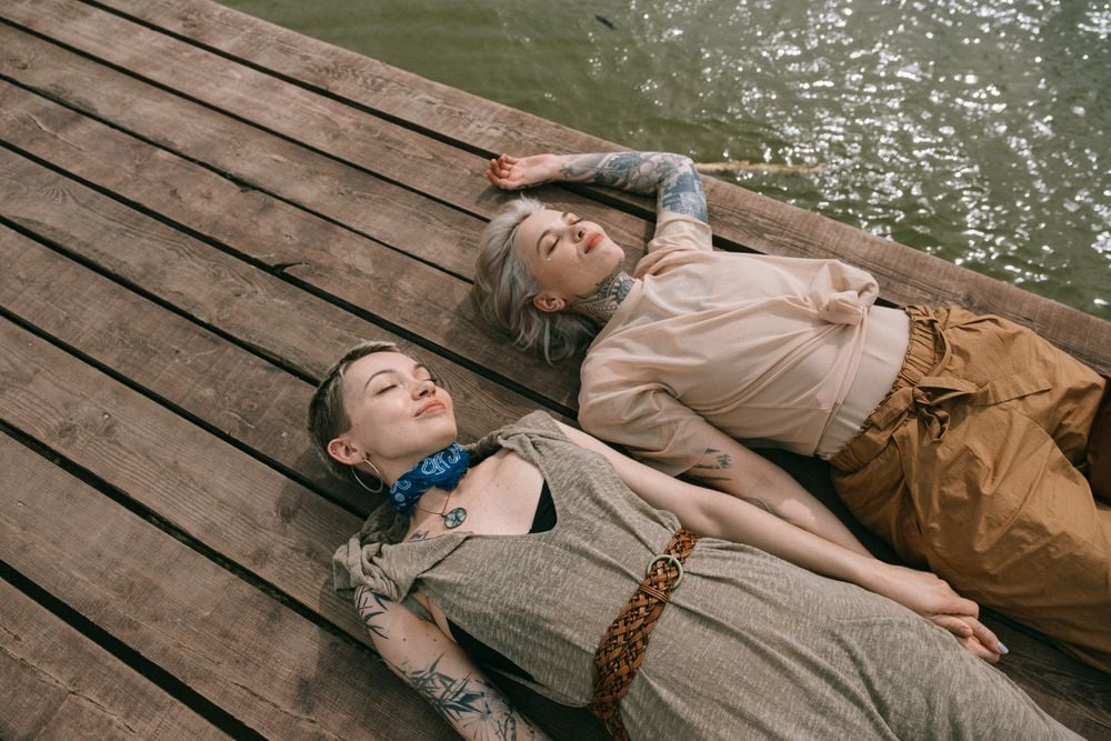 Two people with tattoos lie side by side on a wooden dock near the water, eyes closed and relaxed, enjoying the sunlight.