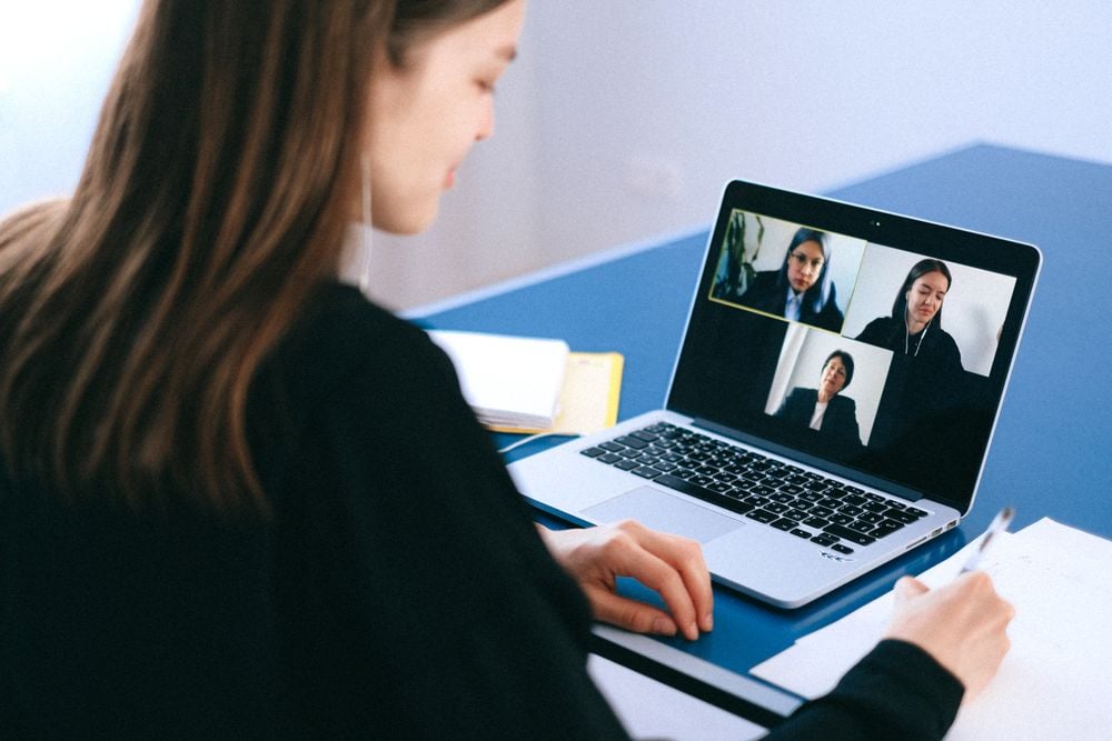 A woman sits at a desk, writing on paper while participating in a video call with three people displayed on her laptop screen.
