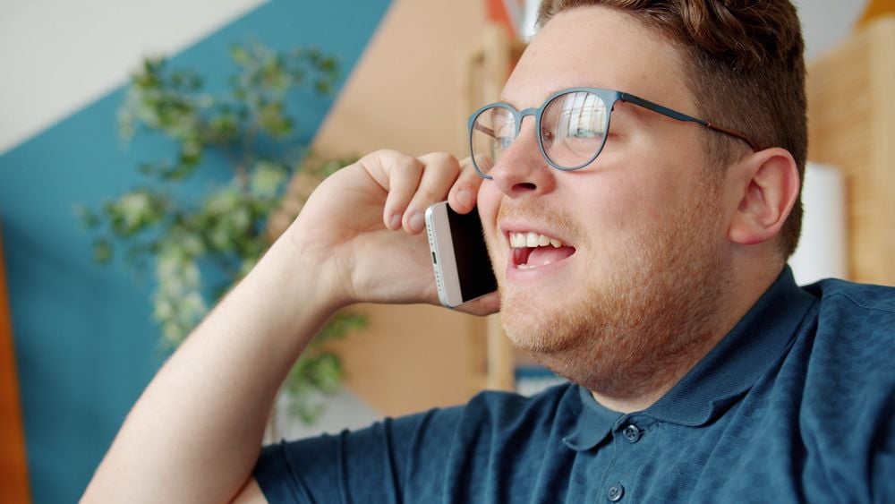 A man with glasses and a beard is smiling while talking on a smartphone indoors. A blurred plant and colorful wall are in the background.