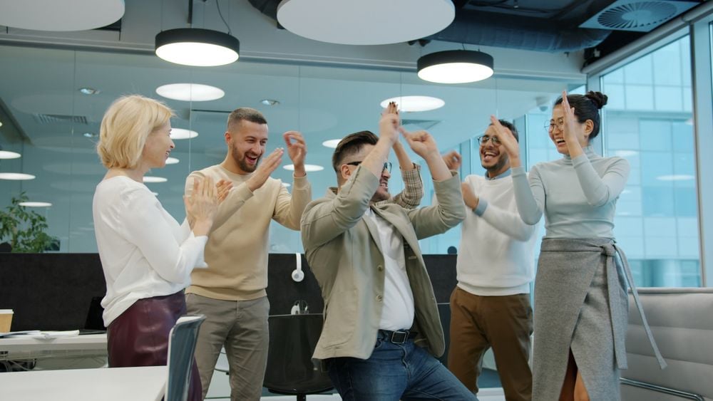 Five people in business attire are celebrating together in a modern office, smiling and raising their hands in excitement. The atmosphere appears energetic and joyful.
