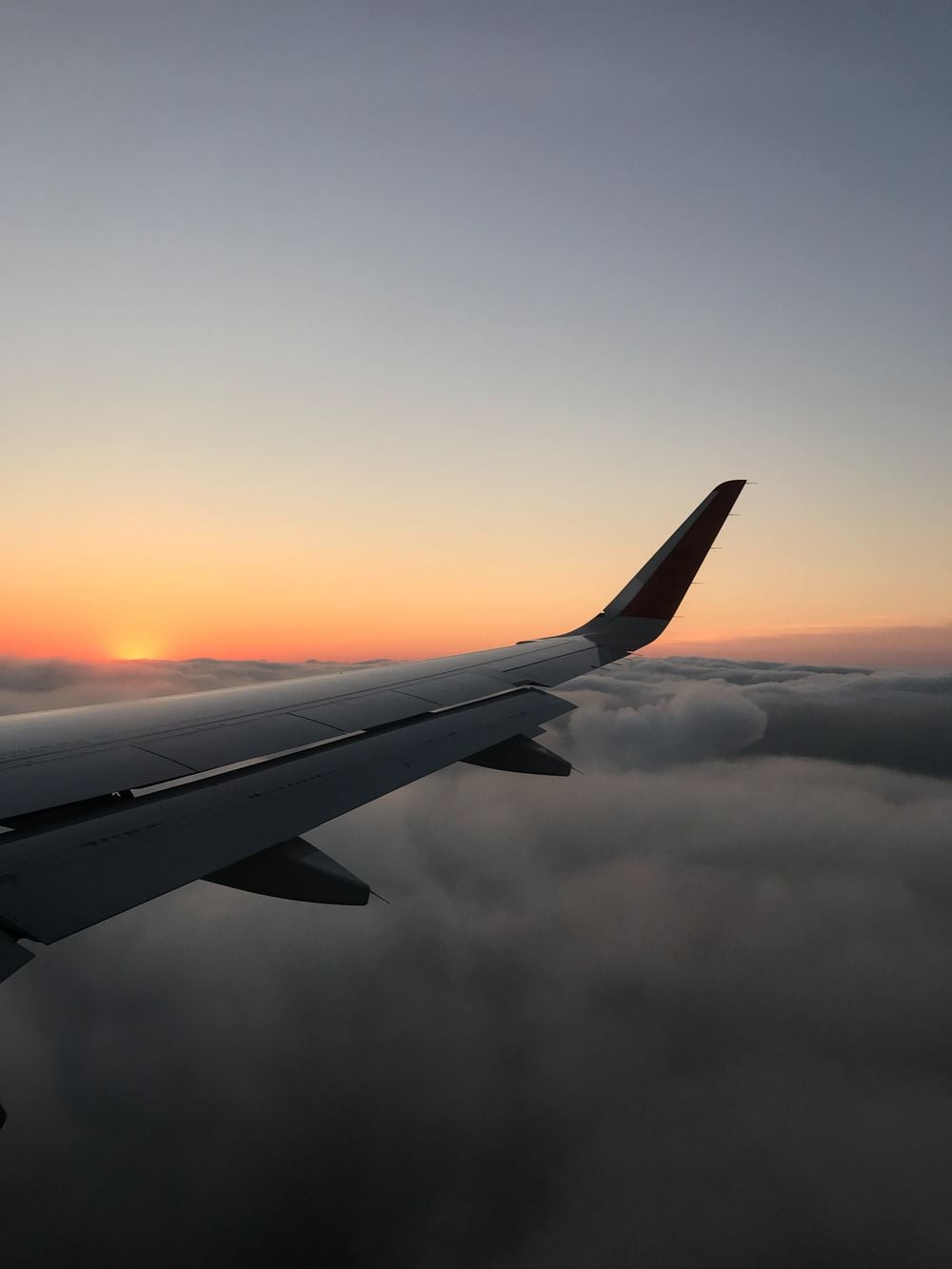 Airplane wing above clouds at sunset, with the sun partially visible on the horizon and a sky blending from orange to blue.