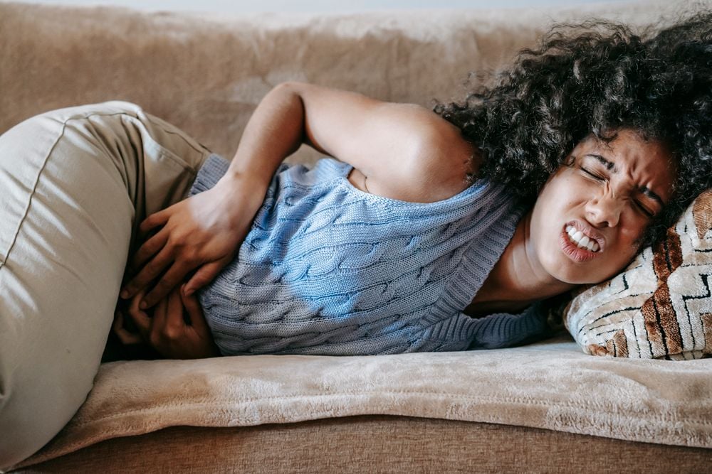 A woman lies on a couch, clutching her stomach in pain, with a pained expression on her face. She wears a blue sweater and beige pants.