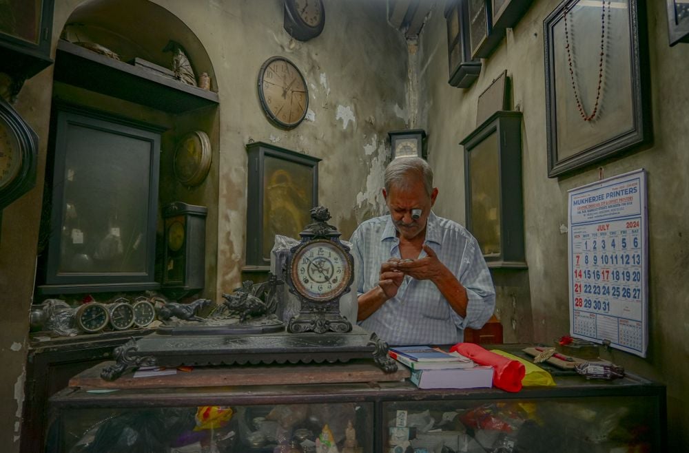 An elderly man repairs a watch in a small, dimly lit workshop filled with antique clocks. A calendar hangs on the wall, and various tools and timepieces are scattered on the counter.