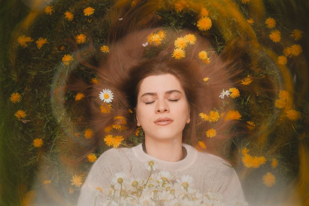 A woman with closed eyes lies on grass, surrounded by yellow and white flowers, with her brown hair fanned out and flowers woven through it, creating a dreamy, peaceful atmosphere.