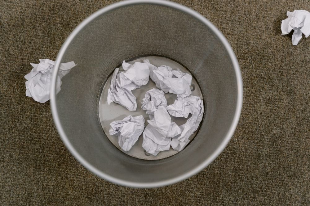 View from above of a metal trash can containing several crumpled pieces of white paper, with more crumpled paper on the carpeted floor around it.