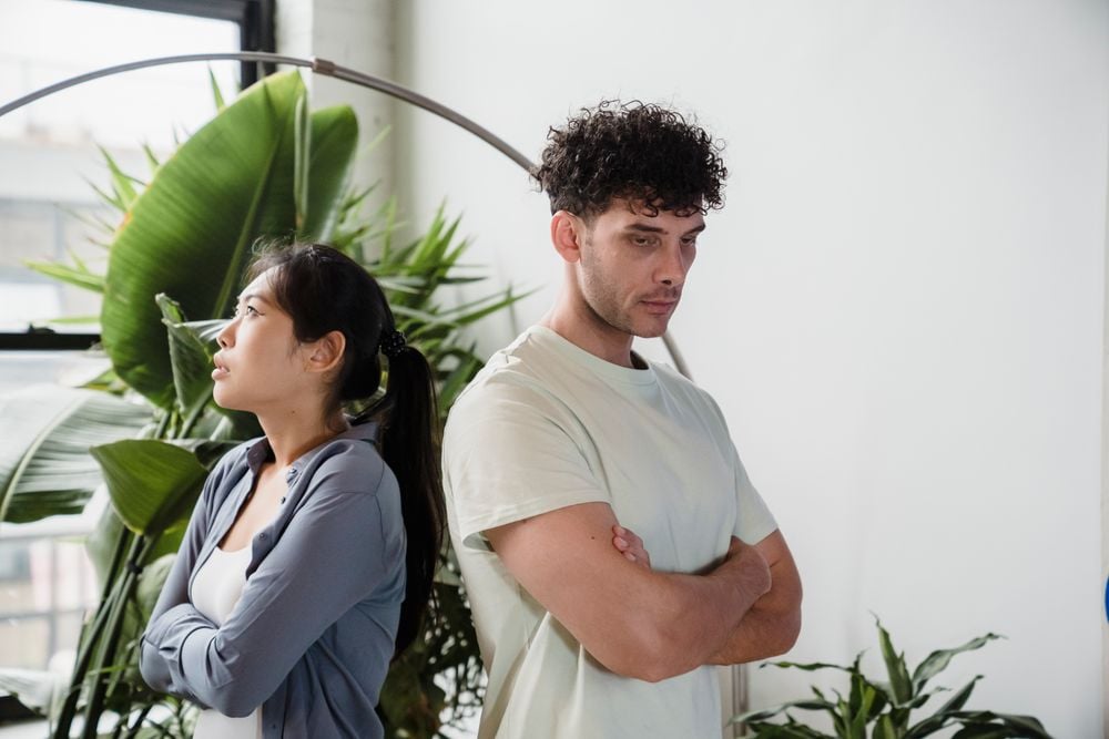 A woman and a man stand back to back with arms crossed, looking away from each other. They appear upset or in disagreement. There are large green plants in the background.