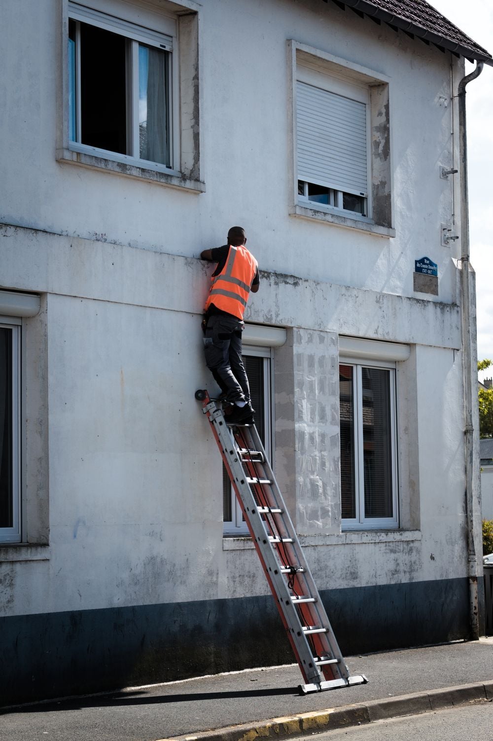 A person in a bright orange safety vest stands near the top of a ladder leaning against a white building, working with their hands near a window on the second floor.