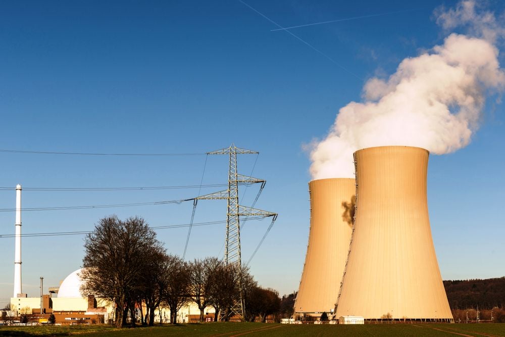 Nuclear power plant with two large cooling towers emitting steam, a tall power line, trees, and clear blue sky in the background.