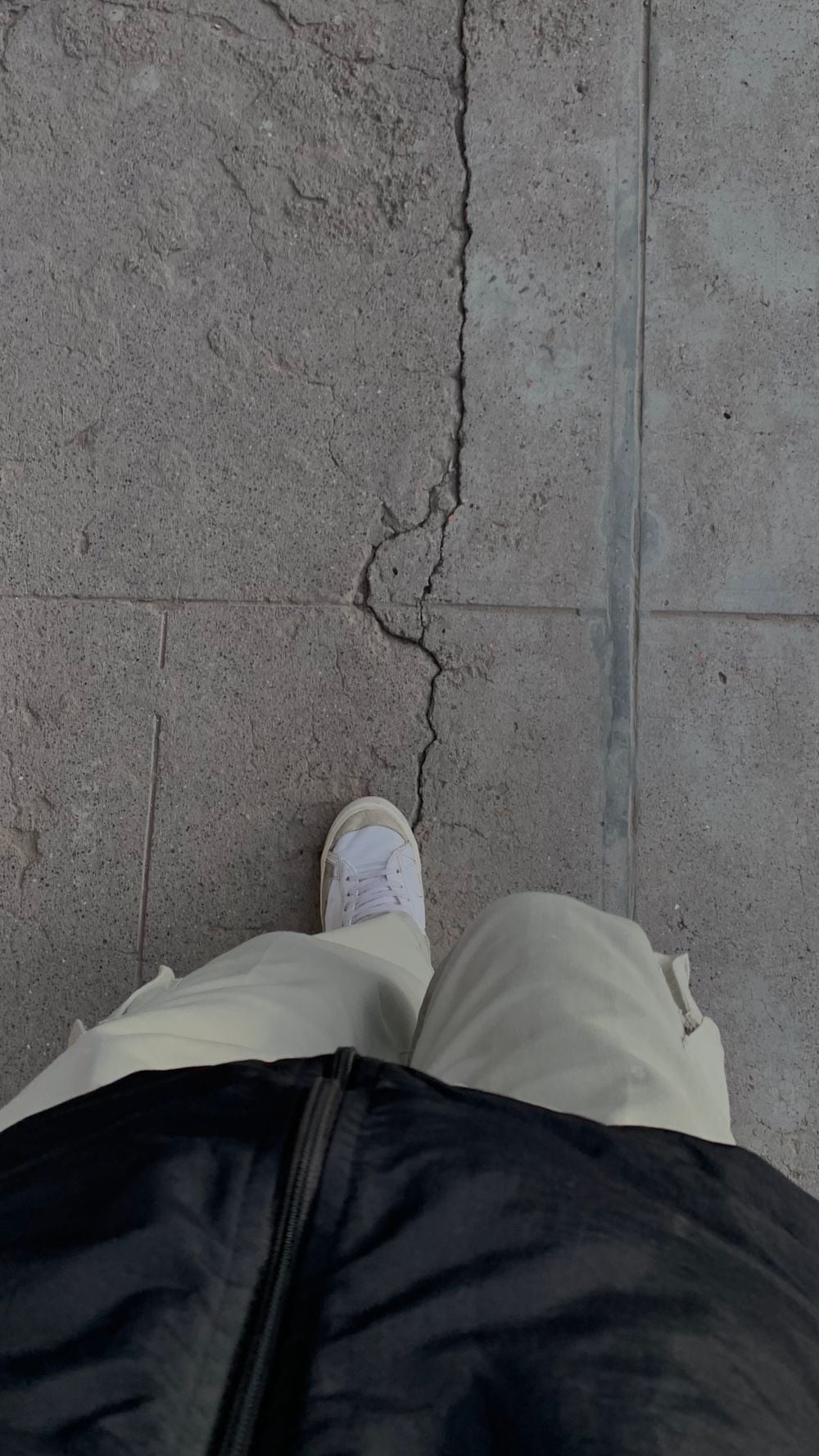 Person wearing white pants and white shoes standing on a cracked concrete sidewalk, partially visible from a top-down perspective. The person is also wearing a dark jacket.
