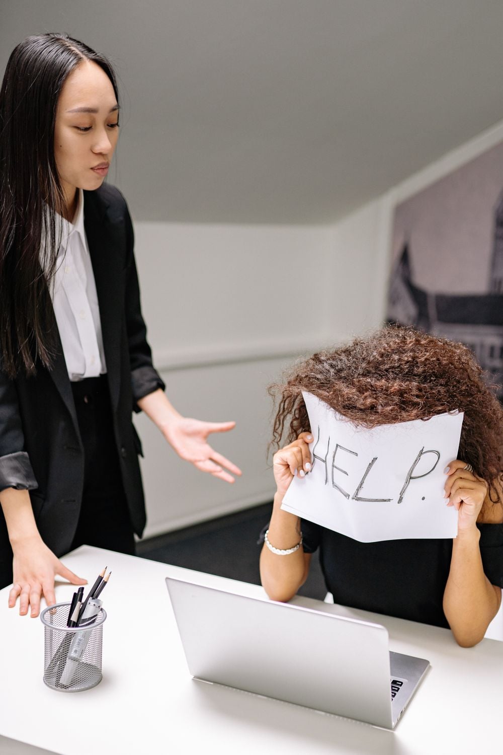 A person sits at a desk holding a paper reading “HELP” over their face, while another person stands beside them, gesturing with concern. A laptop and pencil holder are on the desk.