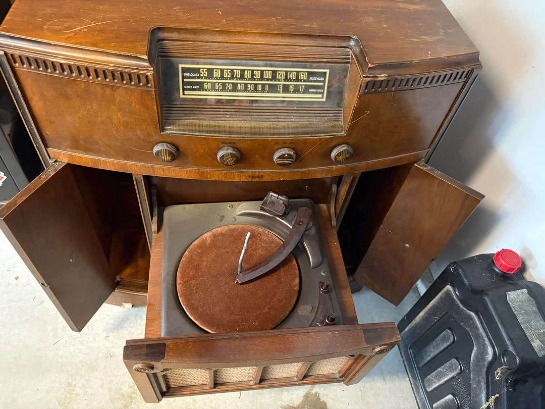 A vintage wooden console radio with an integrated record player, open doors revealing the turntable and speakers, sits on a floor next to a black container.