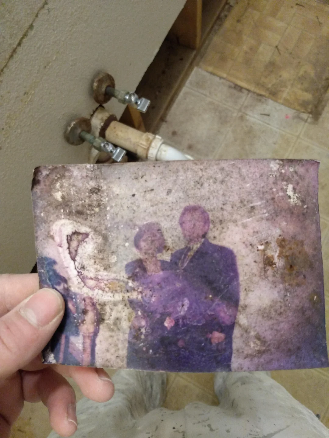 A hand holds a dirty, water-damaged photograph of two people in formal attire. The photo is faded and stained, making the subjects difficult to see clearly. Pipes and a tiled floor are visible in the background.