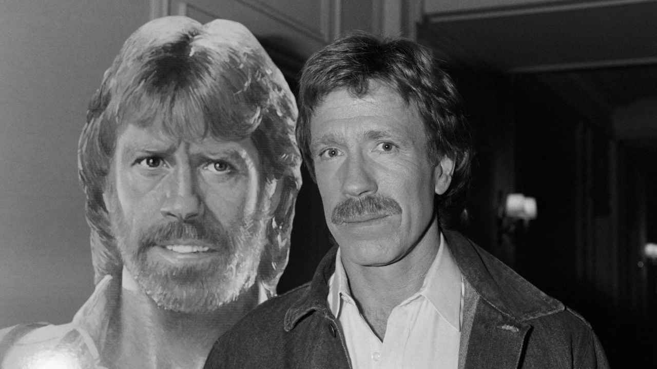 A man with a mustache stands next to a large cutout of himself with longer hair and a beard, both wearing collared shirts; the background is an indoor hallway.