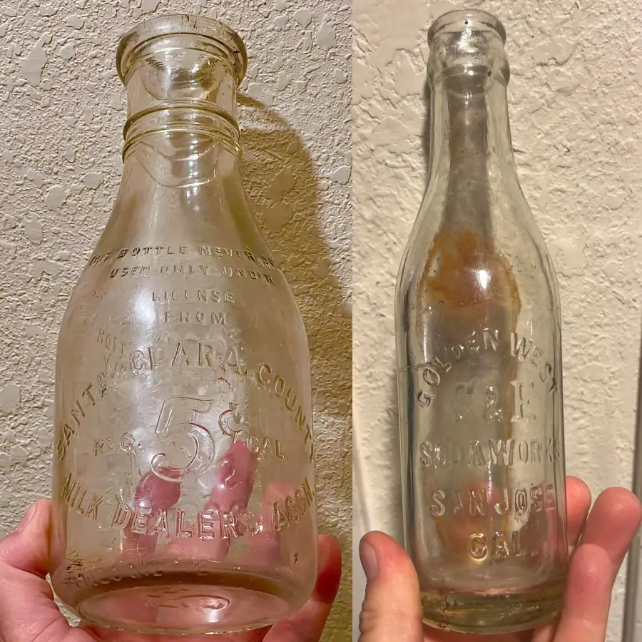 Two old glass bottles held up against a textured wall, one labeled “Santa Clara County Milk Dealers Assoc” and the other “Golden West Soda Works San Jose Cal.” Both have embossed text and show signs of age.