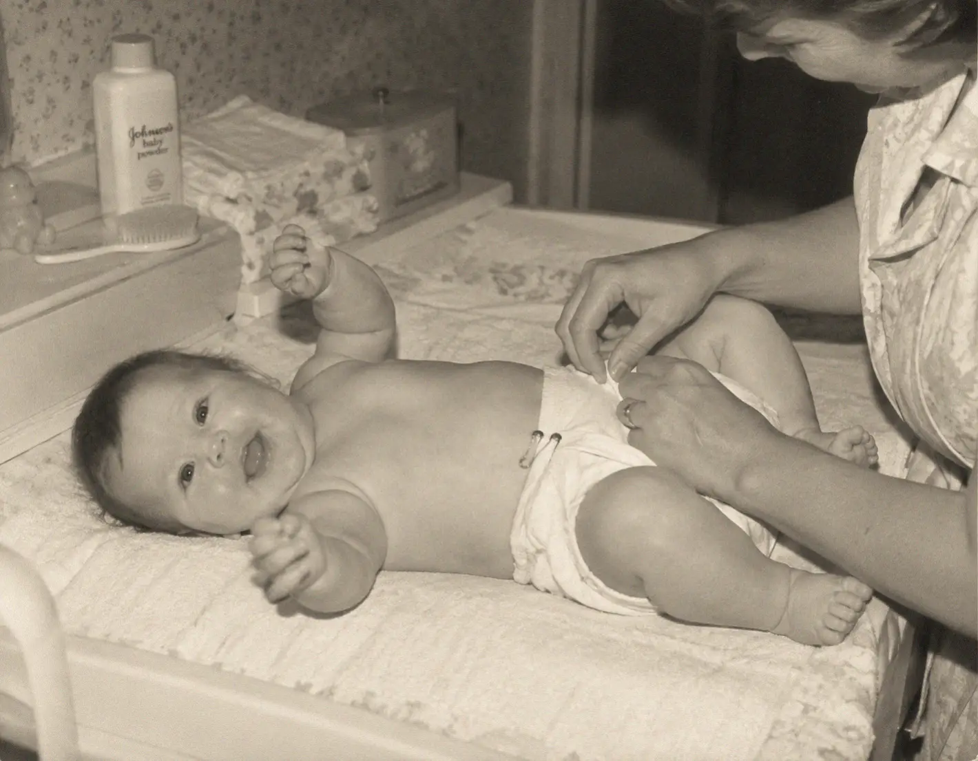 A smiling baby lies on a changing table while an adult fastens a cloth diaper. Baby supplies, including baby powder and a brush, are visible nearby. The scene appears to be from decades ago.